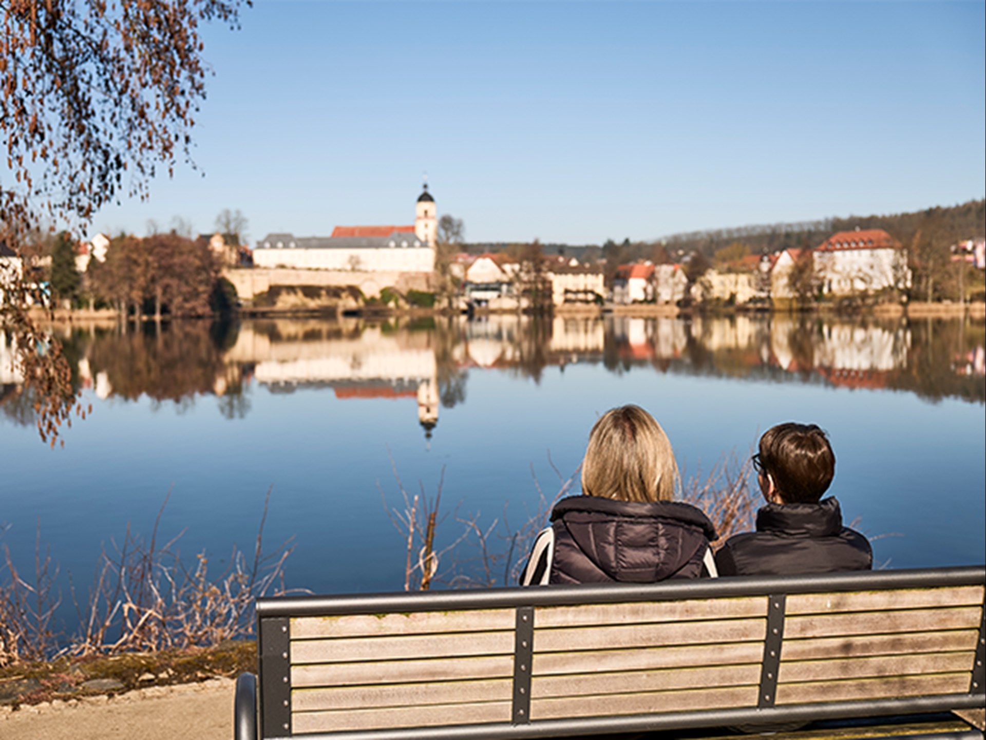 Zwei Frau mit Rückenansicht sitzen auf einer Bank und schauen auf einen großen See