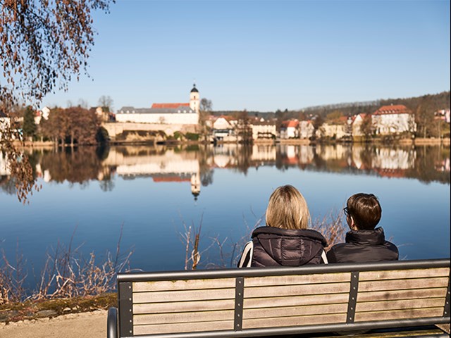 Zwei Frau mit Rückenansicht sitzen auf einer Bank und schauen auf einen großen See